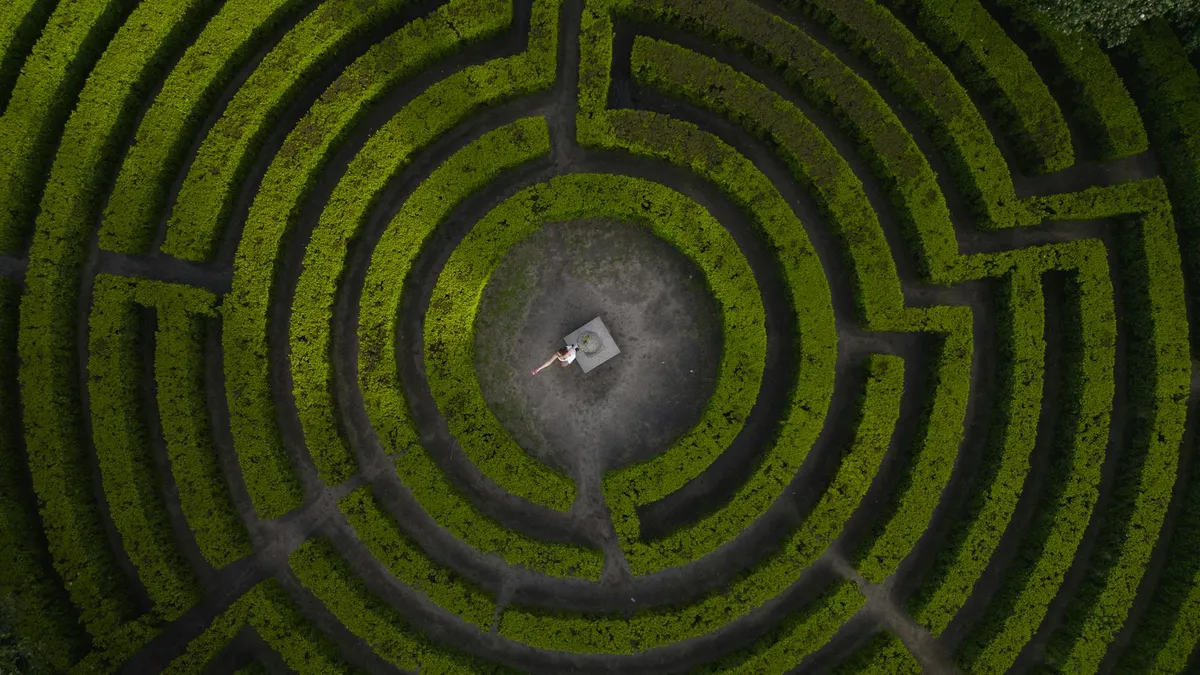 Drone shot of a woman standing in the center of a lush green hedge maze.